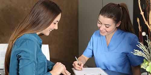 Woman in blue scrubs going over paperwork with patient in a teal shirt with long brown hair